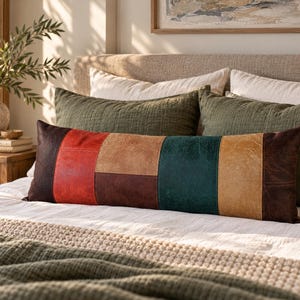 May include: A rectangular lumbar cushion featuring a patchwork design of leather-like panels in dark brown, bright red, tan, and deep teal. The cushion is positioned horizontally on a bed with white bedding and olive green cushions.