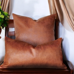 May include: Two brown leather throw pillows with a textured surface. The pillows are stacked on top of each other on a wooden bench.