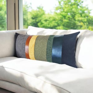 Op de afbeelding: Een lang lumbaalkussen met een gestreept ontwerp. Het kussen heeft vijf strepen in tinten grijs, bruin, geel, groen en blauw. De strepen zijn gemaakt van verschillende materialen, waaronder stof en leer.