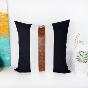 May include: Black and white striped cushion with a brown leather accent strip. The cushion is made of a soft, textured fabric.