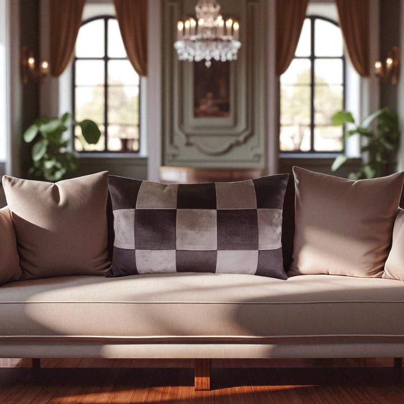 May include: A beige sofa with three pillows. The center pillow is rectangular and has a checkered pattern in shades of brown and beige. The other two pillows are solid beige. The room has large windows and a chandelier.