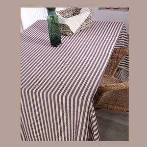 May include: A striped tablecloth with white and burgundy stripes. The tablecloth is laid out on a table with a wicker chair and a green glass vase.