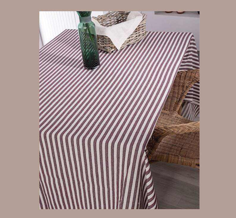May include: A striped tablecloth with white and burgundy stripes. The tablecloth is laid out on a table with a wicker chair and a green glass vase.