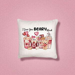 May include: White square pillow with a Valentine's Day theme. The pillow features the phrase "I Love You BEARY Much" above illustrations of teddy bears and coffee cups with heart-shaped balloons and the word "Love".