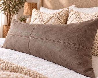May include: A rectangular lumbar pillow in a solid dark brown color with a textured, leather-like finish. The pillow features a prominent horizontal seam running across the center and a vertical seam dividing the left side, creating a paneled look.
