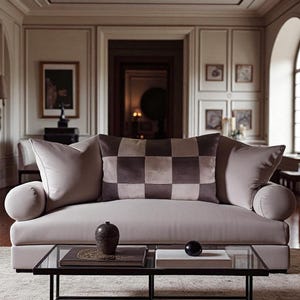May include: A light gray sofa with a checkered pillow and two bolster pillows. A glass coffee table holds a decorative vase, a book, and a black sphere. The room has a neutral color palette with framed artwork on the walls.