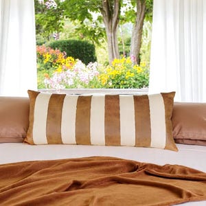 May include: A brown and white striped lumbar pillow on a bed with a brown blanket. The bed is in front of a window with white curtains and a view of a garden.