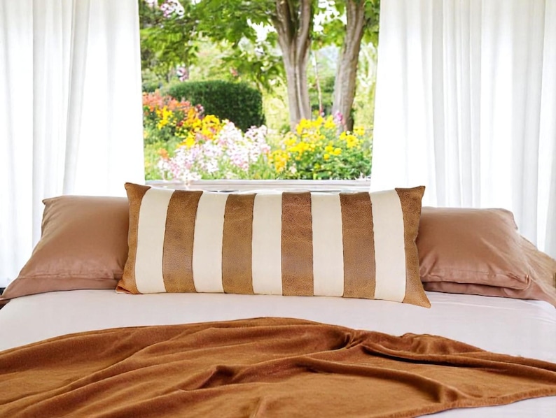 May include: A brown and white striped lumbar pillow on a bed with a brown blanket. The bed is in front of a window with white curtains and a view of a garden.