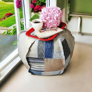 Könnte beinhalten: Ein runder Patchwork-Hocker in neutralen Tönen von Beige, Grau und Blau. Ein rotes Tablett oben hält eine Tasse Kaffee, eine Brille und ein Buch, mit einer rosa Hortensie und einem Fenster im Hintergrund.