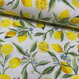 May include: White fabric with a repeating pattern of yellow lemons, lemon slices, green leaves, and small blue flowers. The lemons are in various sizes and orientations, creating a fresh, summery design. The fabric is likely cotton or a similar material.