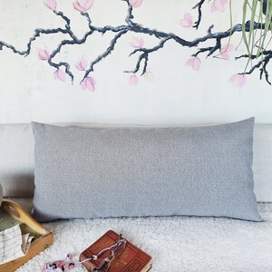 May include: A gray lumbar pillow with a subtle texture sits on a white faux fur throw blanket. The pillow is placed on a couch with a light gray linen fabric. A wooden tray with a checkered pattern is in the foreground.