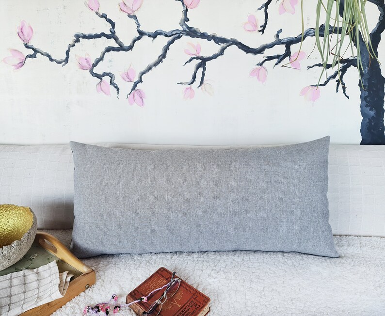 May include: A gray lumbar pillow with a subtle texture sits on a white faux fur throw blanket. The pillow is placed on a couch with a light gray linen fabric. A wooden tray with a checkered pattern is in the foreground.