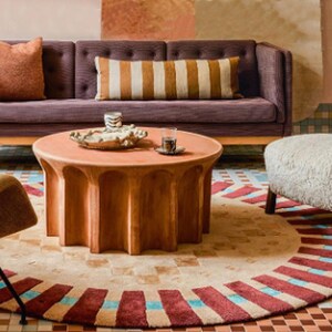 May include: A living room with a purple sofa, a round wooden coffee table, a white sheepskin ottoman, and a patterned rug. The coffee table has a tray with a glass of water and a decorative object.