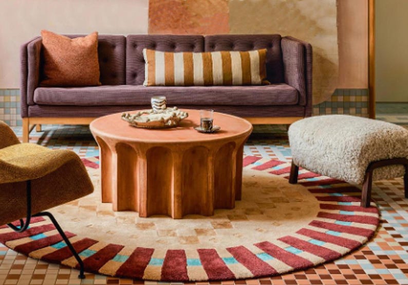 May include: A living room with a purple sofa, a round wooden coffee table, a white sheepskin ottoman, and a patterned rug. The coffee table has a tray with a glass of water and a decorative object.