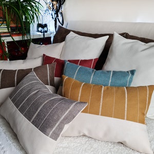 May include: A collection of decorative throw pillows with striped patterns in various colors, including brown, gray, teal, mustard yellow, and red. The pillows are arranged on a white bedspread.