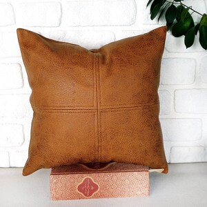 May include: A brown leather pillow with a stitched design. The pillow is sitting on a box with a patterned design.