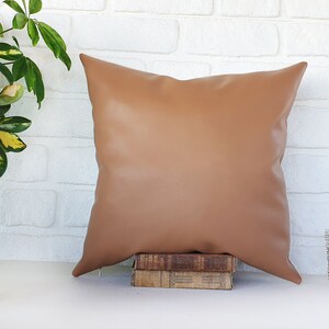 May include: A brown faux leather pillow with a square shape and a smooth surface. The pillow is sitting on top of a stack of books.