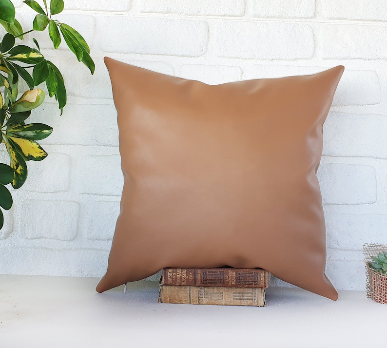 May include: A brown faux leather pillow with a square shape and a smooth surface. The pillow is sitting on top of a stack of books.