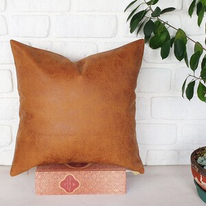 May include: A brown faux leather pillow with a textured surface. The pillow is sitting on a book with a patterned cover.