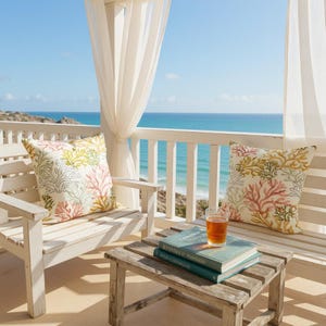 Pu&ograve; includere: Una panca e una sedia in legno bianco con cuscini a motivi corallini si trovano su un patio con vista sull'oceano. Un tavolino di legno contiene libri e un bicchiere di liquido ambrato. Tende bianche trasparenti incorniciano la vista.