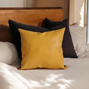 May include: A yellow throw pillow with a smooth, leather-like texture sits on a bed with a white duvet and black pillows.