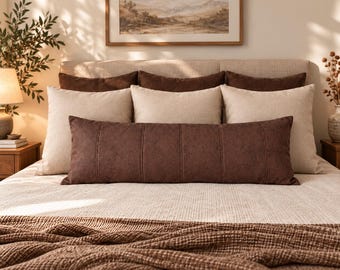 May include: A bed featuring a long rectangular dark brown textured lumbar pillow placed in front of three beige square pillows and three dark brown square pillows. The bed is covered with a light beige textured blanket and a dark brown waffle-knit throw.