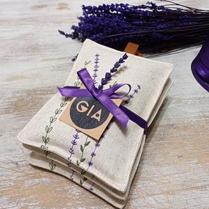 May include: Three white linen sachets with lavender embroidery and a purple ribbon tied around them. A brown tag with the word "GIA" is attached to the ribbon.