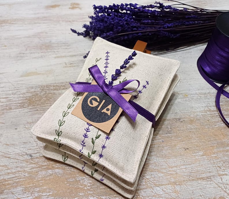 May include: Three white linen sachets with lavender embroidery and a purple ribbon tied around them. A brown tag with the word "GIA" is attached to the ribbon.
