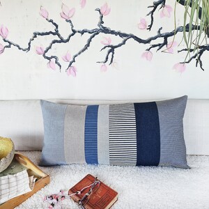 May include: A long rectangular pillow with a patchwork design. The pillow is made of different shades of blue and gray striped fabric. The pillow is sitting on a white couch with a book and a bowl on the surface.
