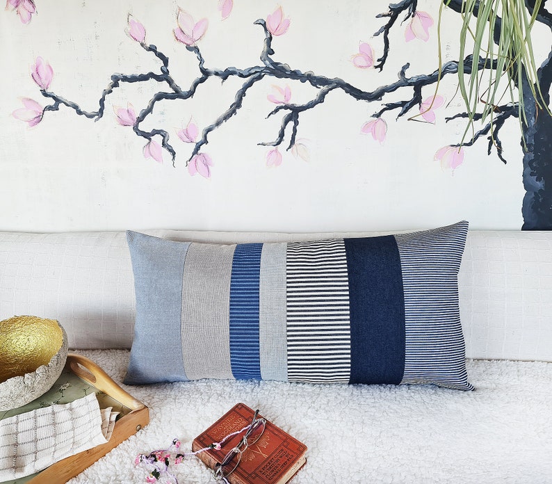 May include: A long rectangular pillow with a patchwork design. The pillow is made of different shades of blue and gray striped fabric. The pillow is sitting on a white couch with a book and a bowl on the surface.
