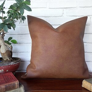 May include: A brown leather pillow with a textured surface. The pillow is sitting on a wooden surface with books.