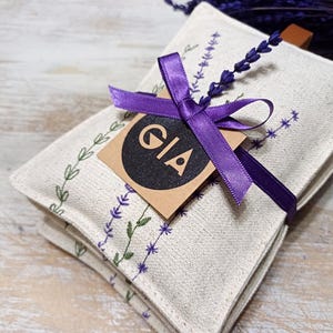 May include: A set of three white linen lavender sachets tied with a purple ribbon and a brown tag that says "GIA".