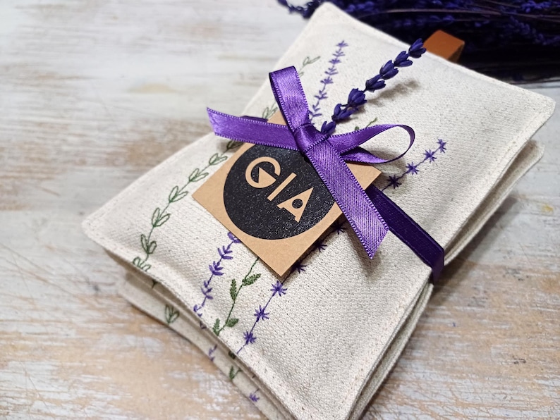 May include: A set of three white linen lavender sachets tied with a purple ribbon and a brown tag that says "GIA".