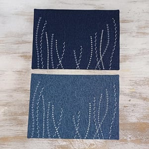 May include: Two blue denim placemats with white embroidered floral designs. The placemats are rectangular and have a subtle texture.