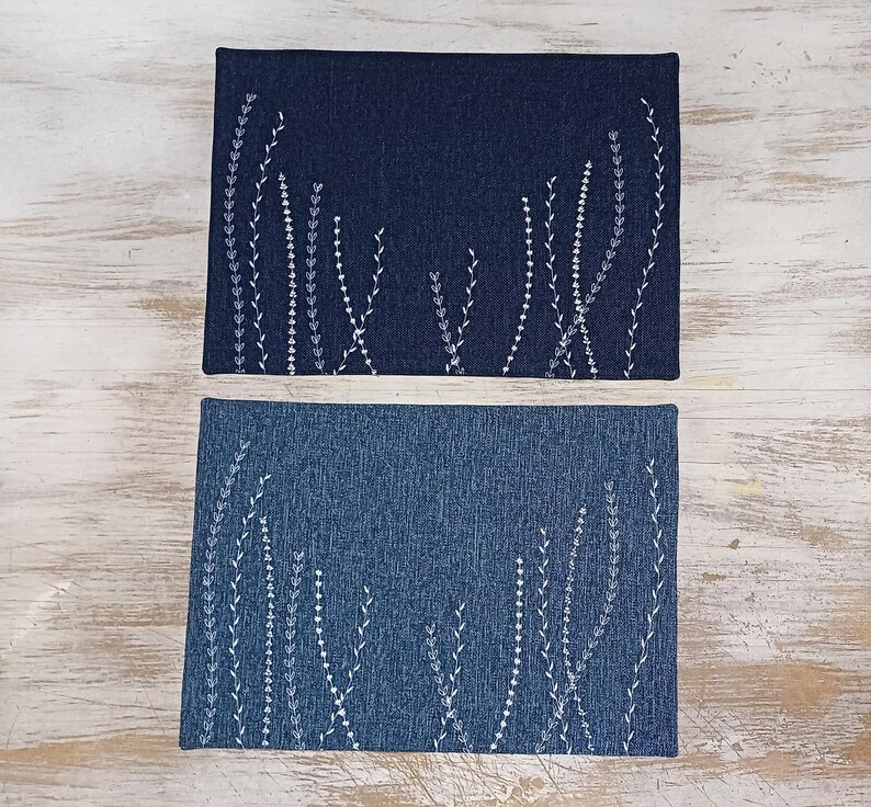 May include: Two blue denim placemats with white embroidered floral designs. The placemats are rectangular and have a subtle texture.