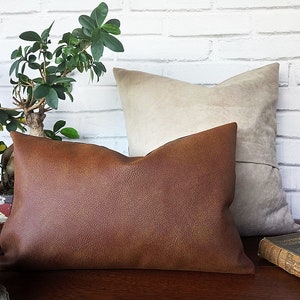 May include: Two decorative throw pillows, one brown leather and one beige suede, on a wooden surface with a book.