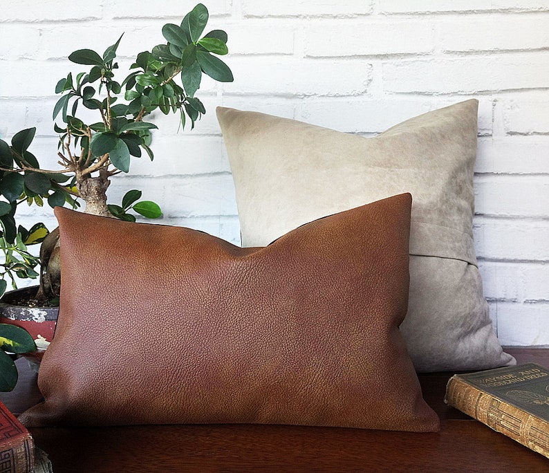May include: Two decorative throw pillows, one brown leather and one beige suede, on a wooden surface with a book.