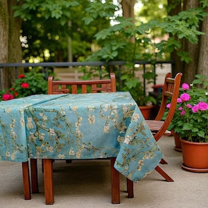Può includere: Una tovaglia quadrata con sfondo azzurro chiaro e motivo floreale, drappeggiata su un tavolo di legno. La tovaglia presenta un motivo di fiori e rami bianchi. Due sedie di legno sono posizionate vicino al tavolo, con piante in vaso sullo sfondo.