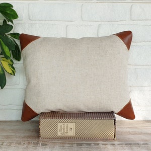 May include: A beige linen pillow with brown leather corners. The pillow is sitting on a wooden surface with a book titled "The Complete Sherlock Holmes" on top of it.
