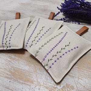 May include: Three beige linen sachets with lavender embroidery. The sachets have brown leather ties and are filled with dried lavender.