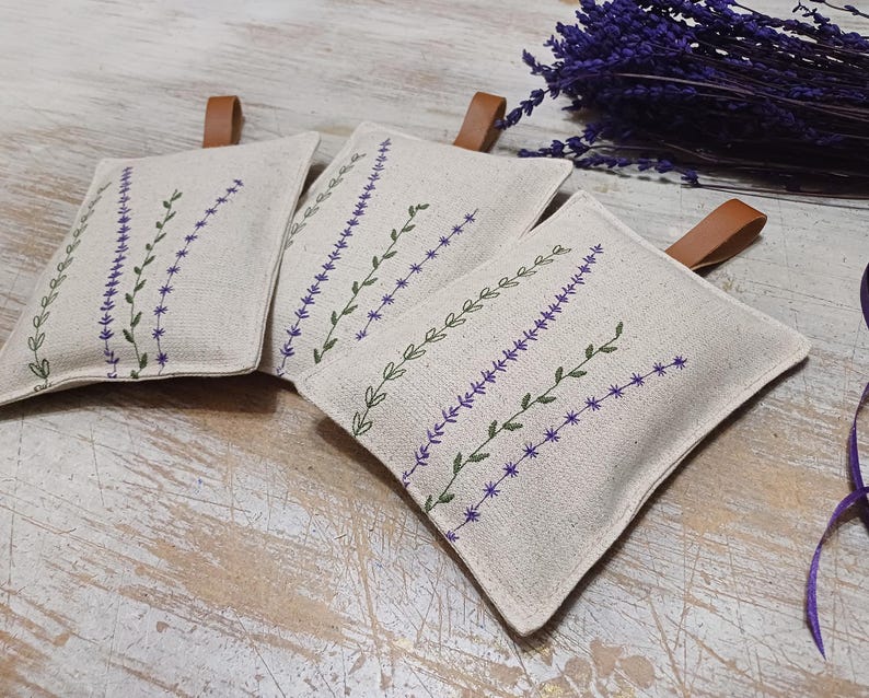May include: Three beige linen sachets with lavender embroidery. The sachets have brown leather ties and are filled with dried lavender.