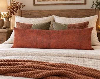 May include: A long, rectangular lumbar pillow in a deep rust-orange color with a subtle, textured, dotted pattern and vertical stitched seams. It is positioned horizontally across the center of a bed layered with cream-colored bedding.