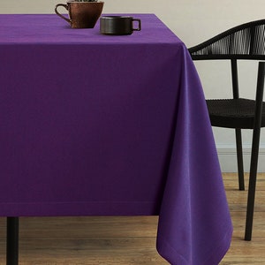 Op de afbeelding: Een paarse tafelkleed gedrapeerd over een tafel met een zwarte stoel op de achtergrond. Het tafelkleed heeft een gladde, gelijkmatige textuur en hangt tot op de grond.