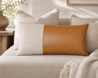 May include: A rectangular throw pillow featuring a color-blocked design with one half made of a light beige textured fabric and the other half made of a smooth tan faux leather material. The pillow is positioned horizontally on a light-colored sofa.