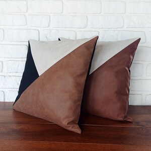 May include: Two decorative throw pillows with a geometric design. Each pillow features a brown faux leather triangle, a beige linen triangle, and a black triangle. The pillows are placed on a wooden surface.