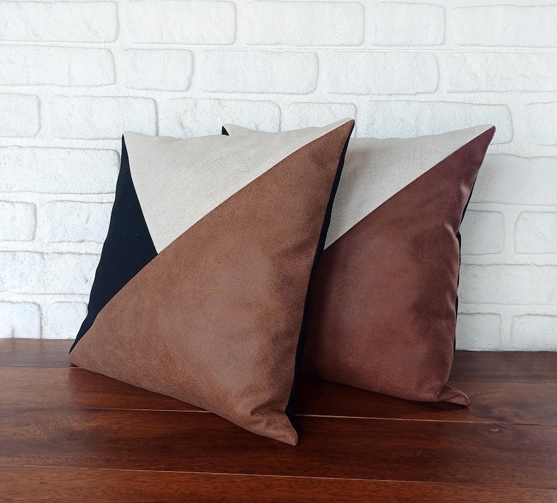 May include: Two decorative throw pillows with a geometric design. Each pillow features a brown faux leather triangle, a beige linen triangle, and a black triangle. The pillows are placed on a wooden surface.
