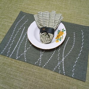 May include: A green placemat with a white embroidered design of branches. A white plate with a floral pattern and a green and white checked napkin with a black leather napkin ring is on top of the placemat.