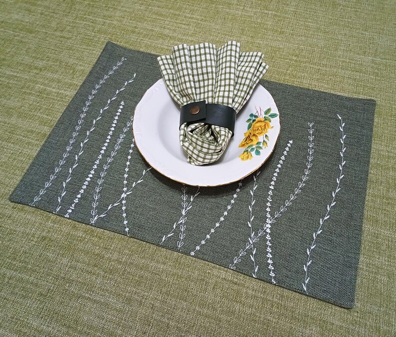 May include: A green placemat with a white embroidered design of branches. A white plate with a floral pattern and a green and white checked napkin with a black leather napkin ring is on top of the placemat.