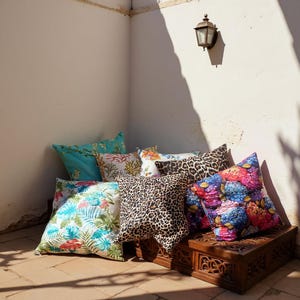 Pu&ograve; includere: Una collezione di cuscini decorativi con vari motivi, tra cui floreali, corallo e leopardati. I cuscini sono disposti su un baule di legno in un angolo soleggiato, creando un'atmosfera accogliente.