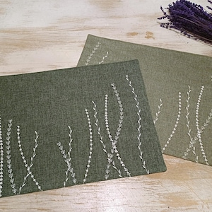 May include: Two green placemats with white embroidered floral designs. The placemats are made of a textured fabric.